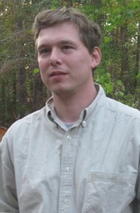 A man in a tan shirt standing in a wooded area.