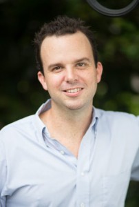 A man in a blue shirt smiling in front of a tree.