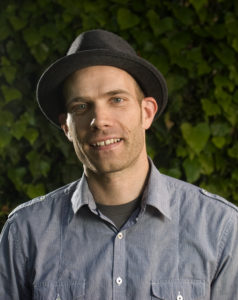 A man in a hat smiling in front of bushes.