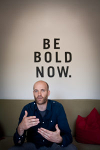 A man sitting on a couch with the words be bold now.