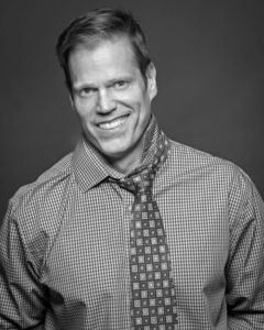 A black and white photo of a smiling man wearing a checkered shirt and tie.