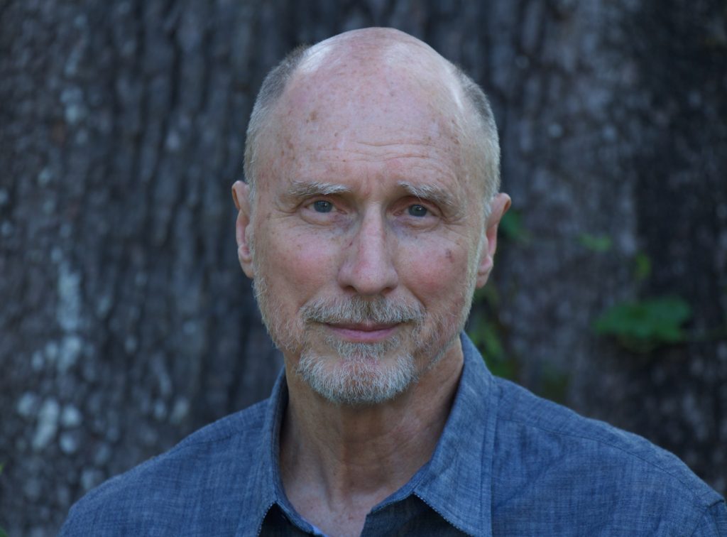 A bald man in a blue shirt standing in front of a tree.